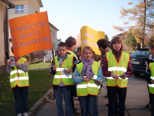 Uczniowie ubrani w odblaskowe kamizeli i trzymający dwa transparenty przed szkołą.
